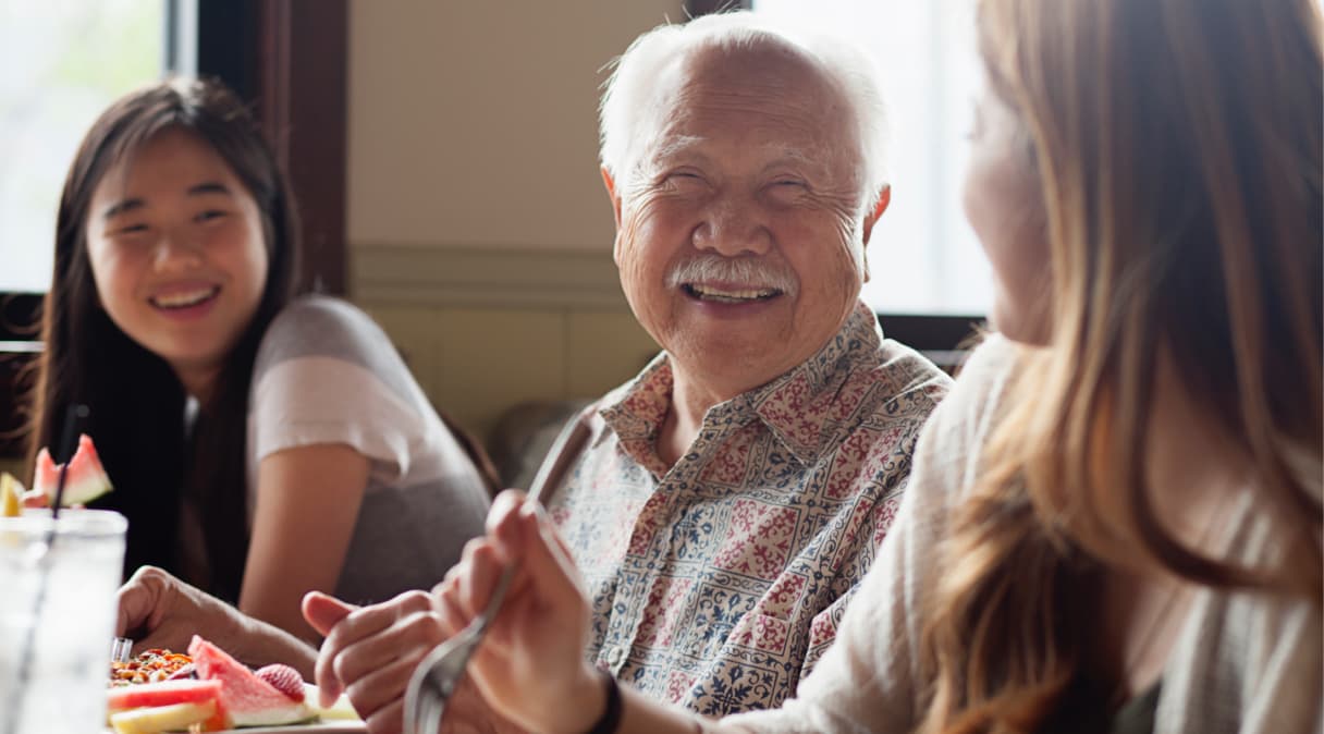 A group of three people in a dining setting sit and smile at each other 