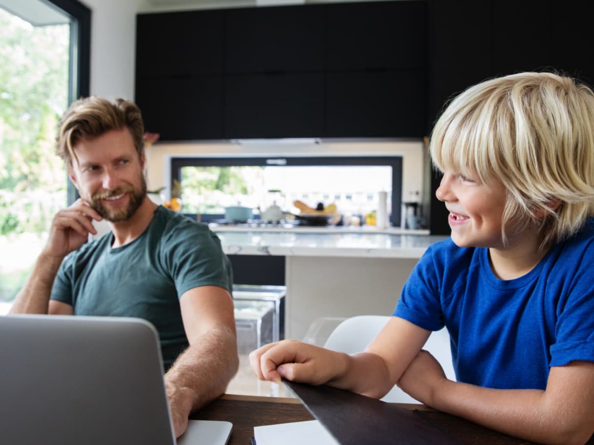 A father figure smiling across the table at a young boy next to multiple digital screens.