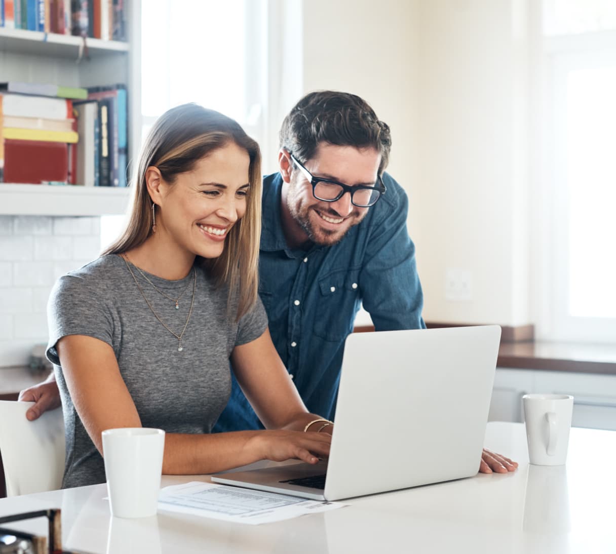 A man and woman sit in front of a laptop at a desk in a home office setting. 