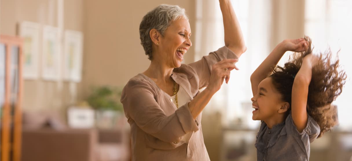 A middle-aged woman and a child smiling at each other, with hands in the air, in a residential setting.